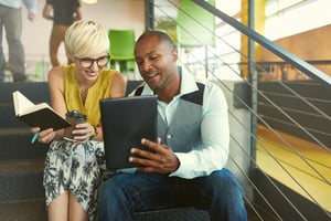 Two creative millenial small business owners working on social media strategy using a digital tablet while sitting in staircase-1